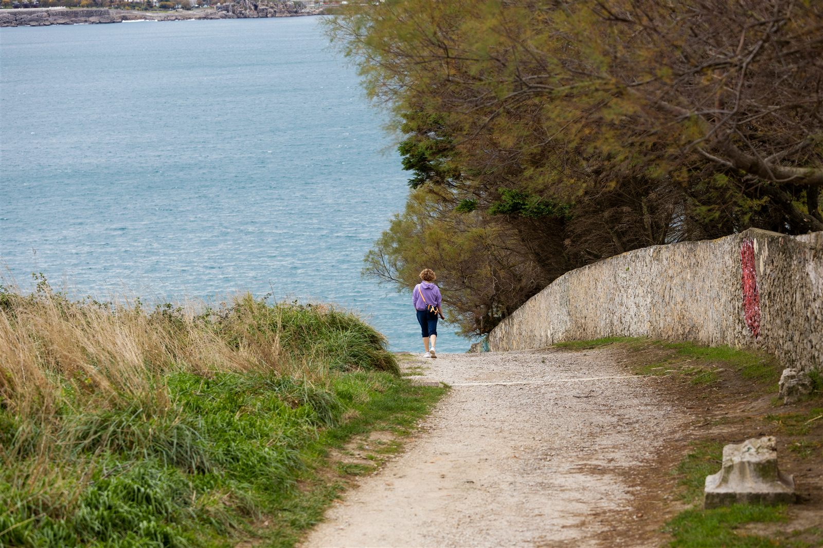Photo of a woman walking away along a gravel coastal path beside the sea, seen from behind