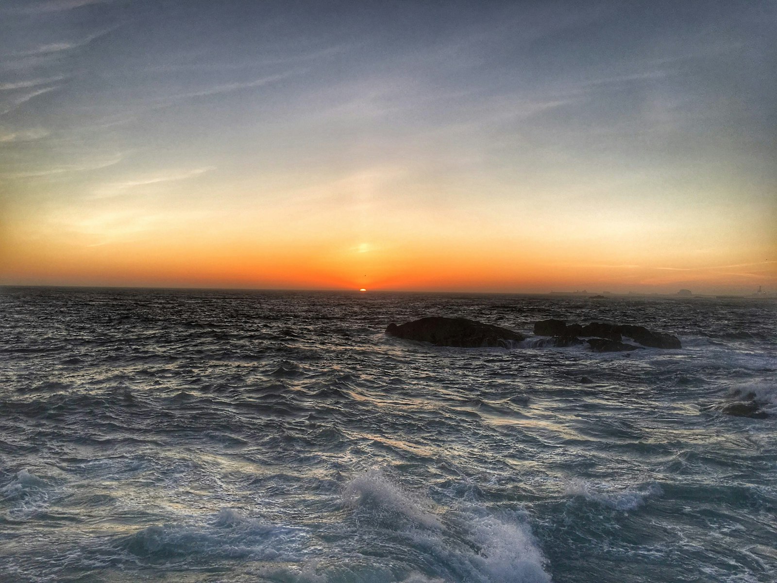 Photo of the sun setting low over the ocean, orange sky reflecting on rolling waves near dark rocks