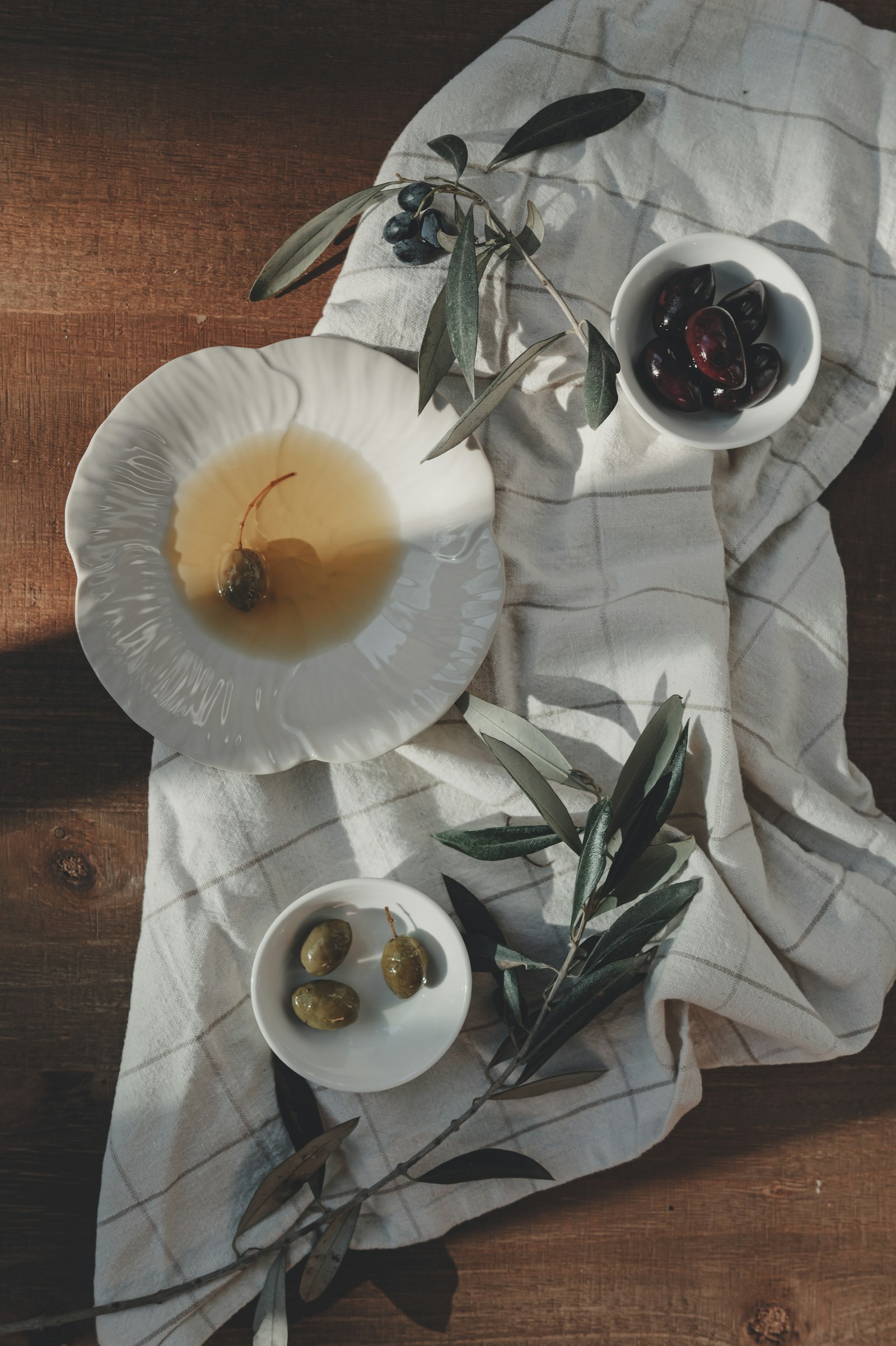 Overhead photo of green and black olives in small ceramic bowls, olive oil in a shallow dish, and olive branches on a linen cloth over a wooden table